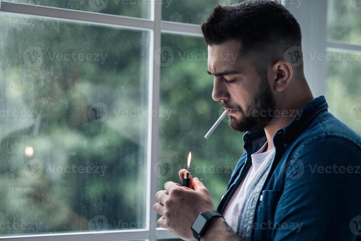 Urban lifestyle, emotional portrait of young handsome man sad and depressed, smoking. Unhappy feeling bittered suffer some problem. Bearded man lights cigarette with lighter, on window background photo