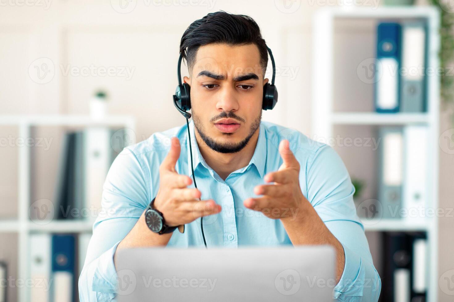 Concerned young Arab man in headset making online call with business partner, solving problem at office. Upset millennial guy having virtual meeting, webcam conference on laptop photo