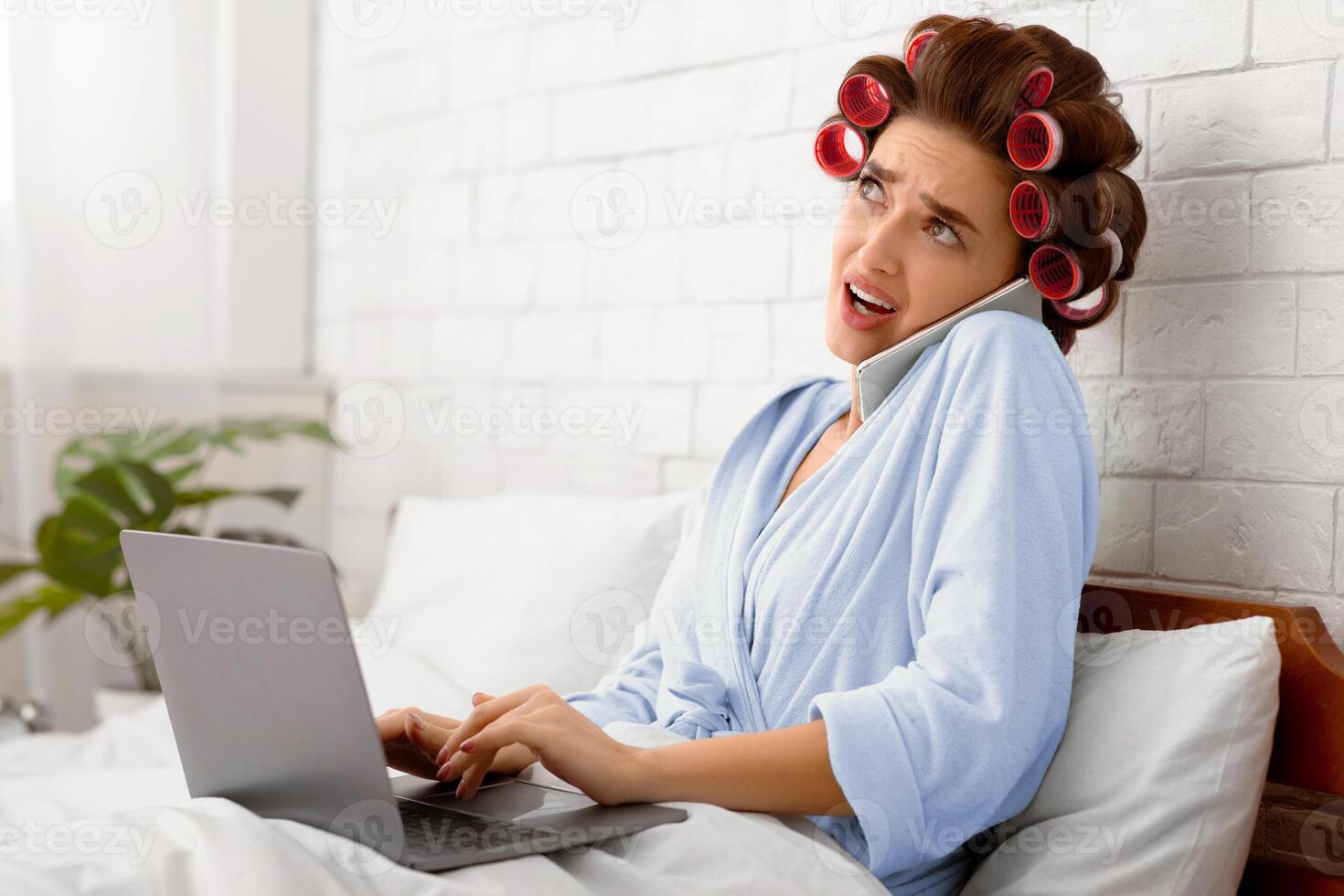 Confused Housewife Talking On Mobile Phone Using Laptop Computer Working Online Sitting In Bed In Modern Bedroom At Home. Mobile Communication, Distance Job And Freelance Work. Side View photo
