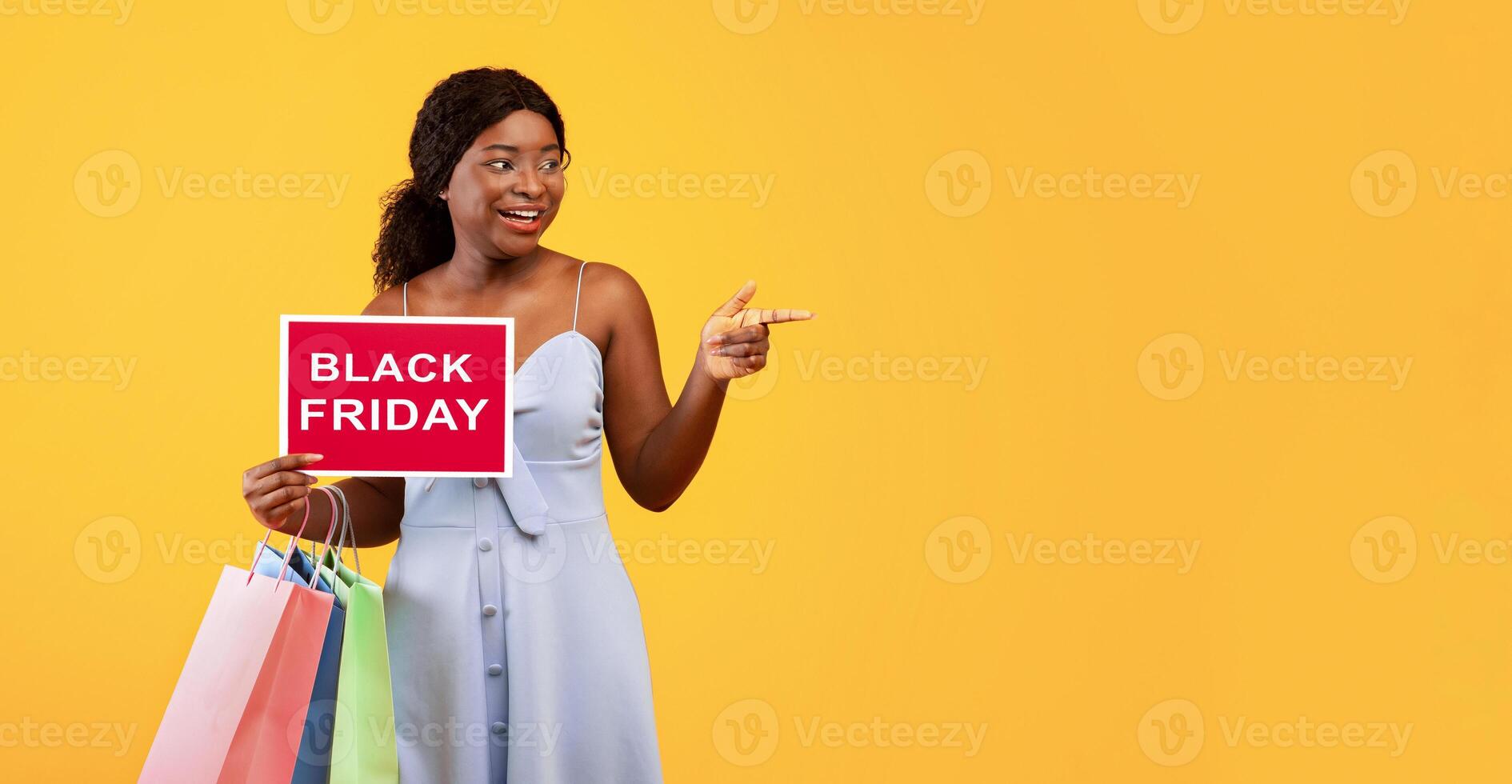Charming young black woman with shopper bags holding BLACK FRIDAY sign and pointing aside at empty space, orange studio background. African American lady having special offer for sale day photo