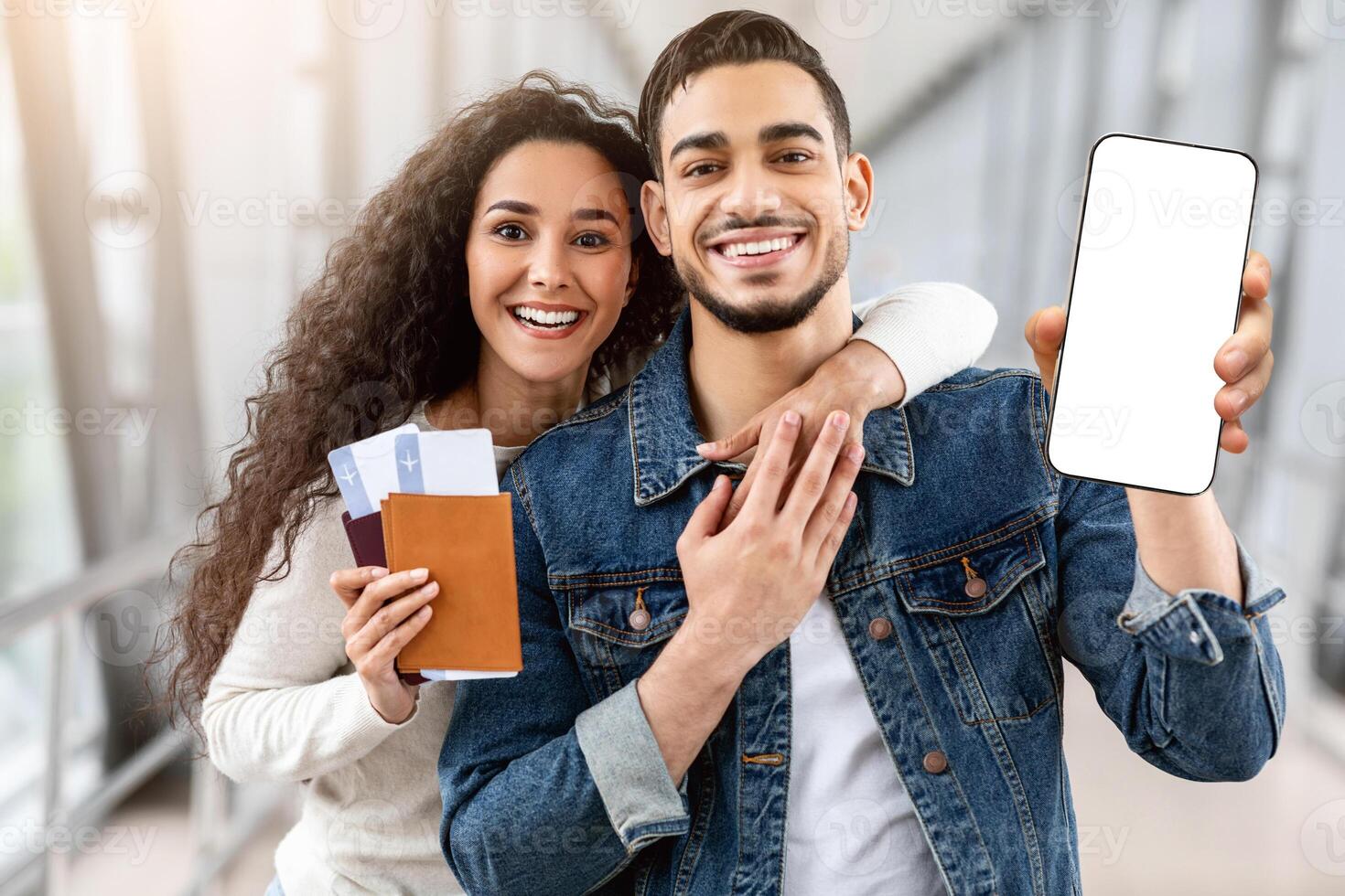 Best Booking App. Happy Arab Couple Holding Passports With Tickets And Blank Smartphone While Posing Together At Airport, Middle Eastern Spouses Recommending New Mobile Application, Mockup photo