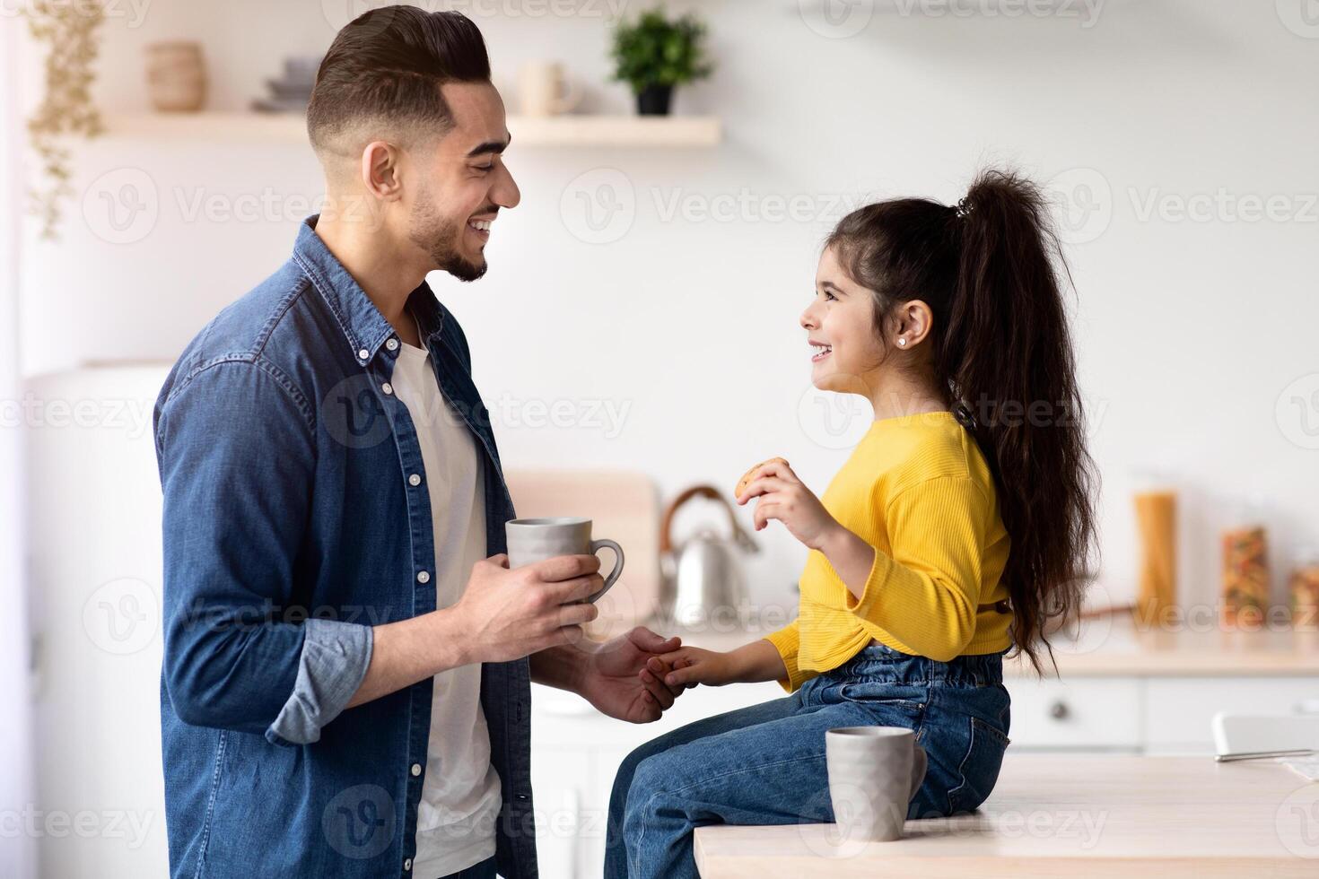 Snack Time. Cute Little Girl And Her Arab Dad Having A Bite In Kitchen At Home Together, Cheerful Middle Eastern Father And His Adorable Female Child Drinking Cocoa And Eating Cookies, Side View photo