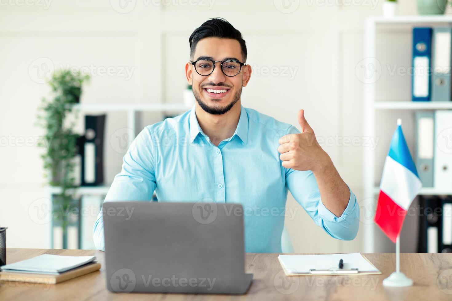 Online French lesson. Foreign languages tutor teaching class on web, using laptop pc, showing thumb up at home office. Positive Arab guy professor giving lecture, speaking to pupils on internet photo