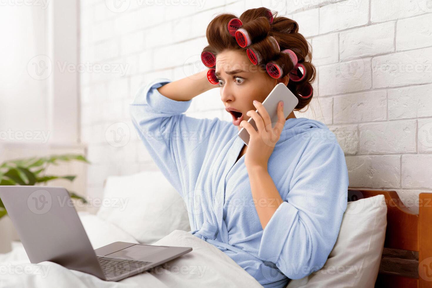 Shocked Housewife Talking On Cellphone Using Laptop Computer Reading Shocking News Online Sitting In Bed Touching Head, Curling Hair With Curlers In Modern Bedroom At Home. Side View Shot photo