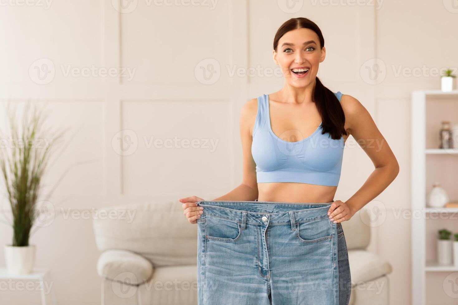 Successful Weight Loss. Thin Girl Comparing Old Oversize Jeans After Dieting Standing Indoors. Copy Space photo