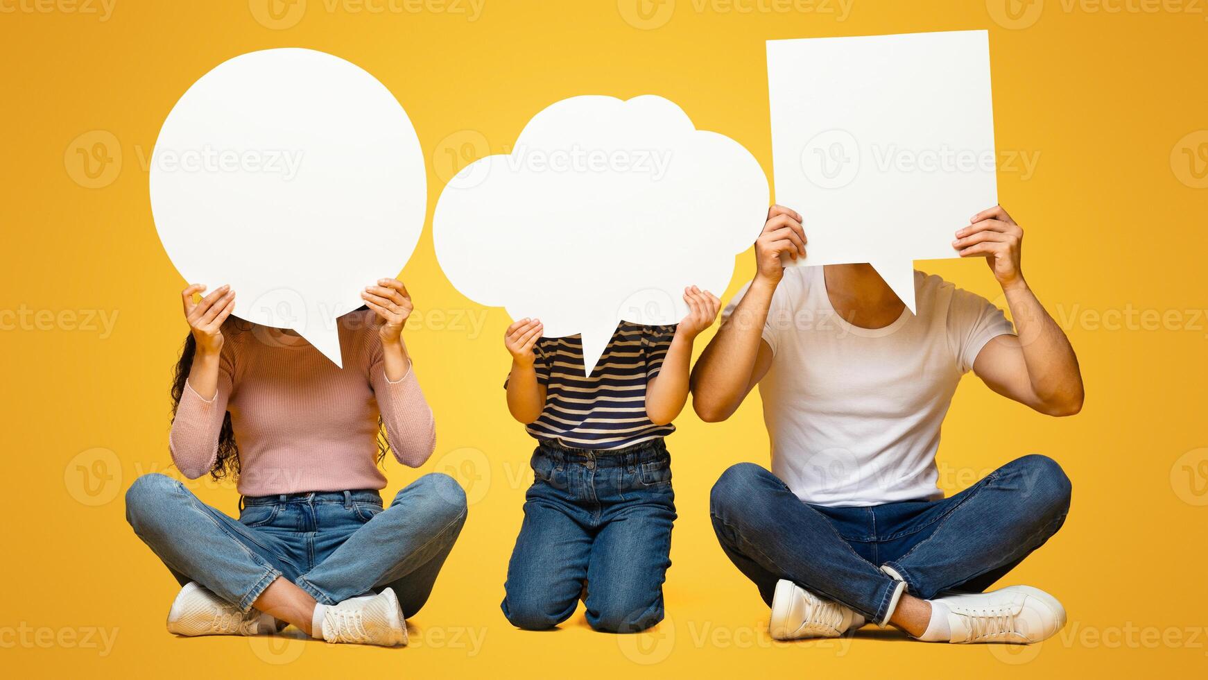 Diverse Thoughts. Father, Mother And Little Daughter Holding Different Empty Speech Bubbles In Front Of Their Faces, Unrecognizable Family Sitting On Floor On Yellow Studio Background, Copy Space photo