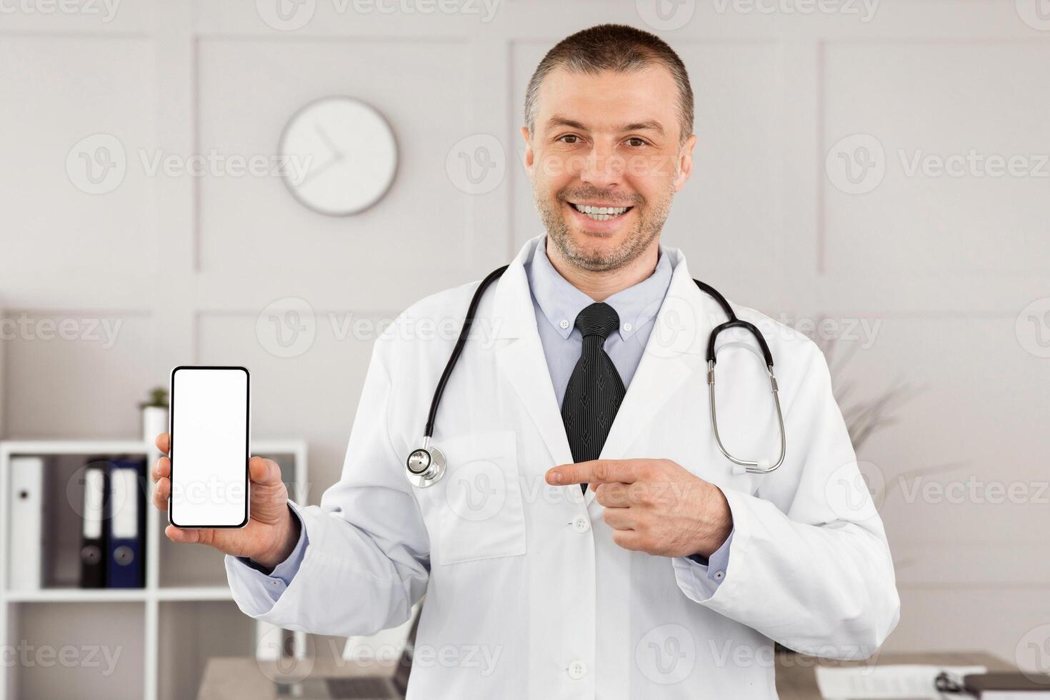 Telemedicine Concept. Portrait of mature male medical practitioner holding smartphone with white blank screen for mockup template, pointing at gadget and smiling, wearing unifrom and stethoscope photo