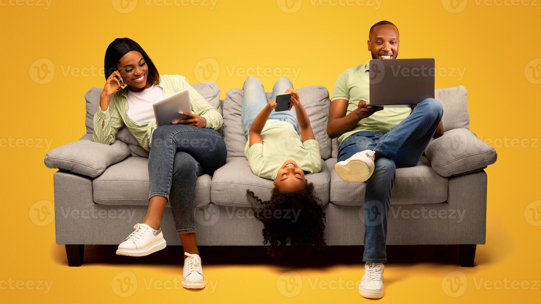 Gadgets addiction. Young black family of three holding and using different electronic devices while sitting on sofa on yellow background. Parents and their daughter with modern gadgets photo