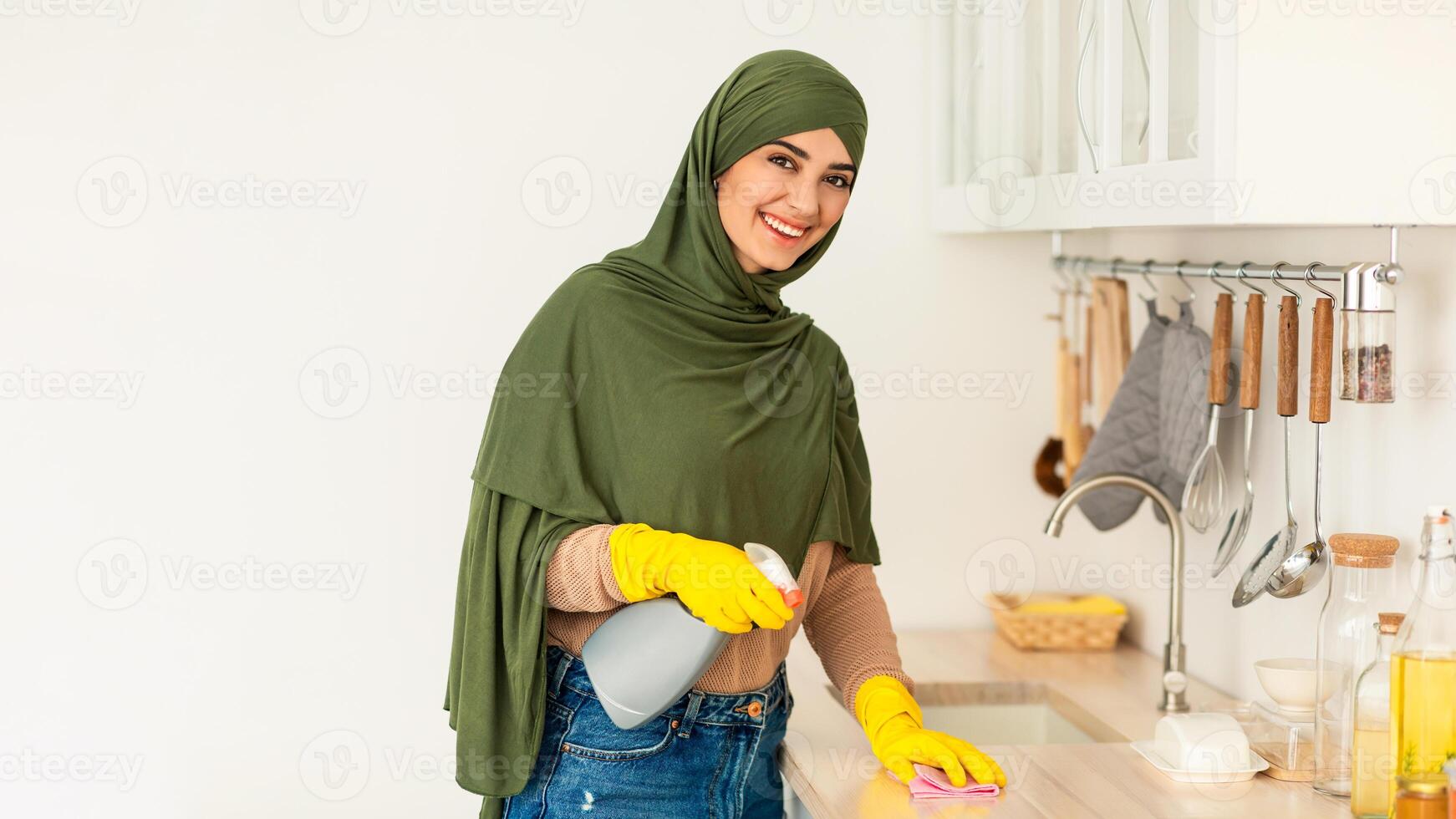 Portrait of happy islamic housewife in hijab and protecitve yellow rubber gloves cleaning countertop at kitchen. Positive lady keeping her home tidy, wiping surface with cloth looking at camera photo