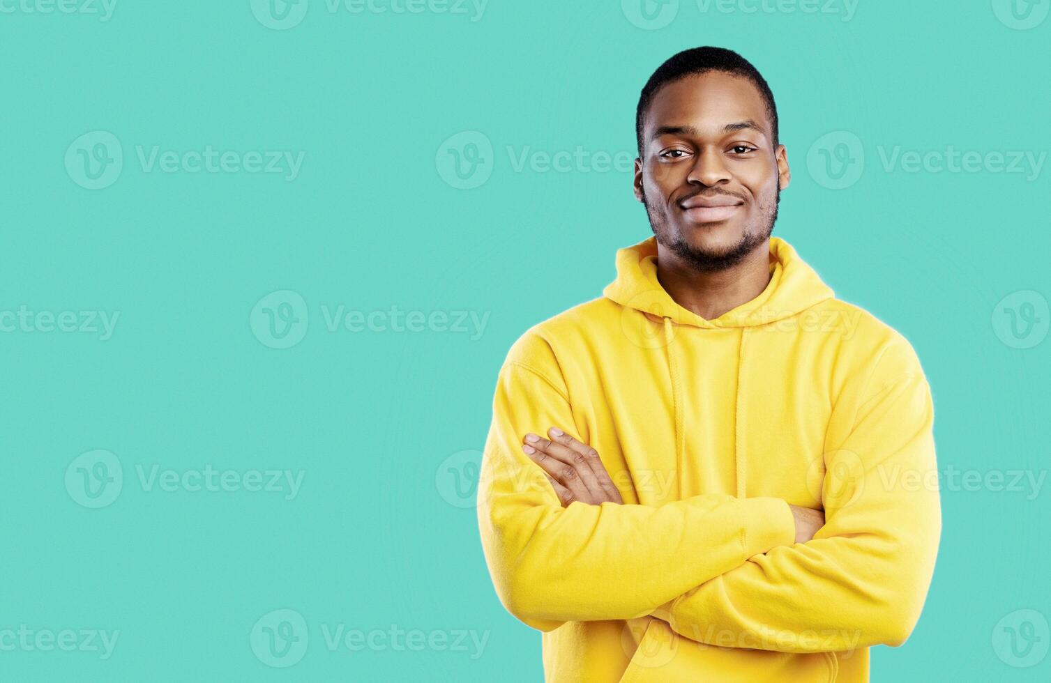 Handsome Black Millennial Man Crossing Hands Posing Smiling To Camera Standing In Studio On Blue Background, Wearing Yellow Hoodie. Shot Of Confident African American Guy photo
