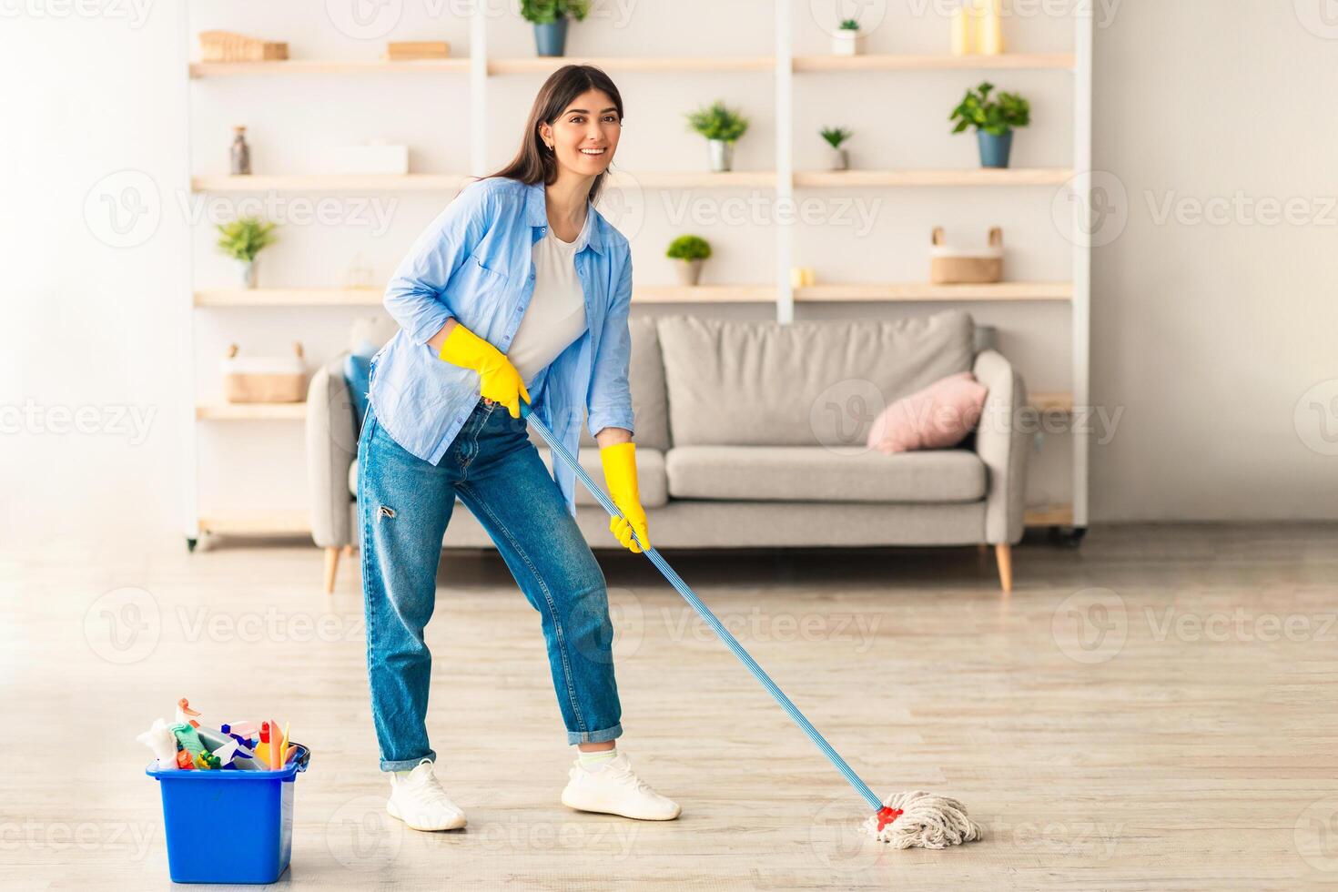 Chores Concept. Portrait of casual woman washing hardwood laminate flooring using string mop pad, standing near bucket with cleaning agents and detergents, posing and looking at camera photo