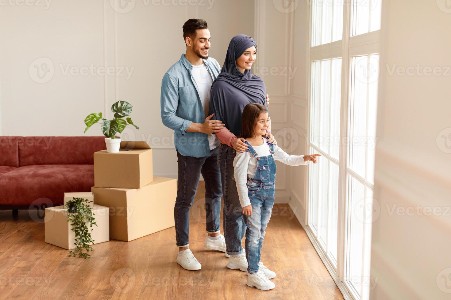 Full Body Length Portrait Of Excited Muslim Family Looking Out And Showing View From Window In Their New Apartment. Renting And House Move Concept. Smiling Little Girl Pointing At Glass photo