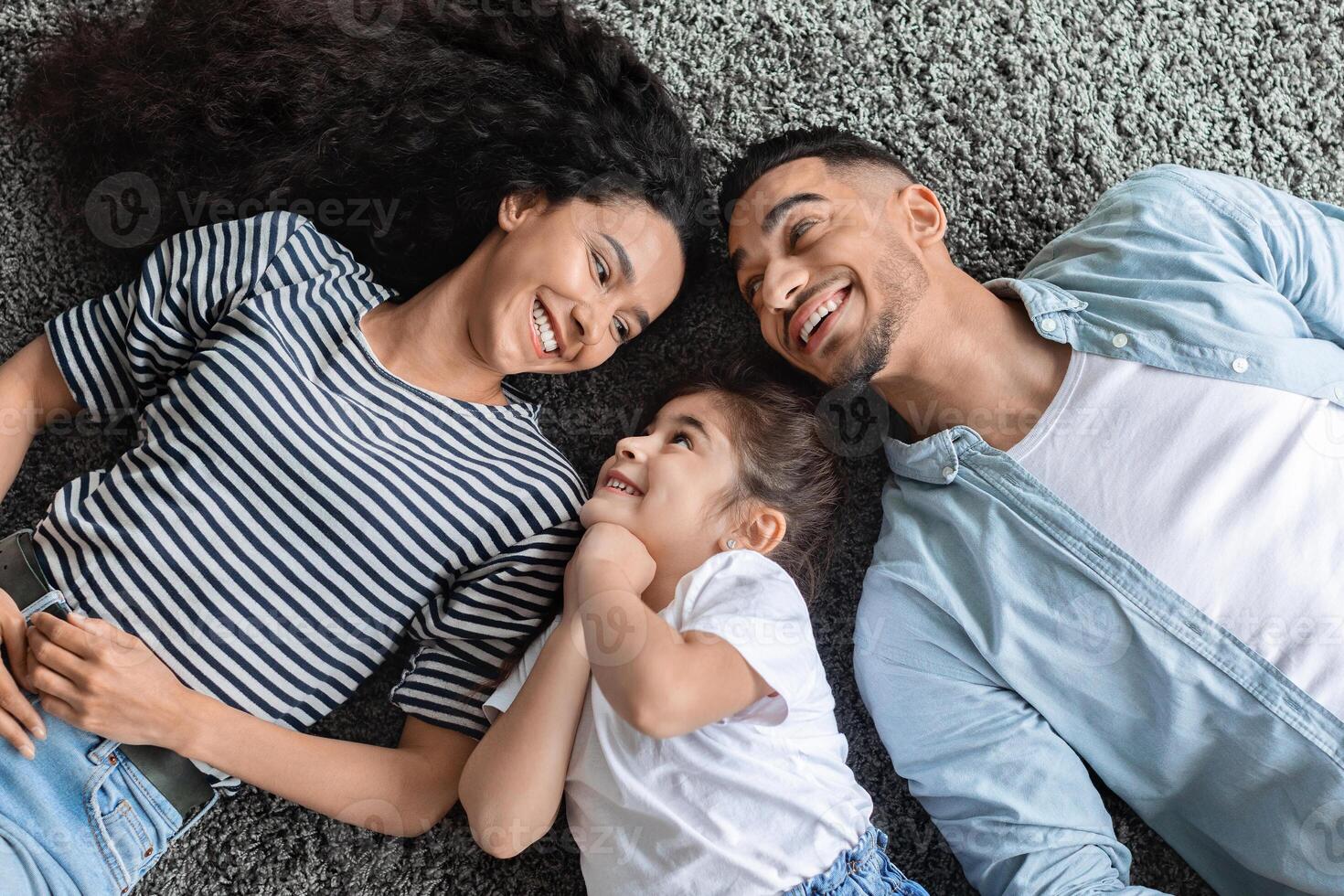 Cheerful arabic family father, mother and preschool daughter laying on floor and laughing, enjoying time together at home. Middle eastern parents spending weekend with their kid, top view photo