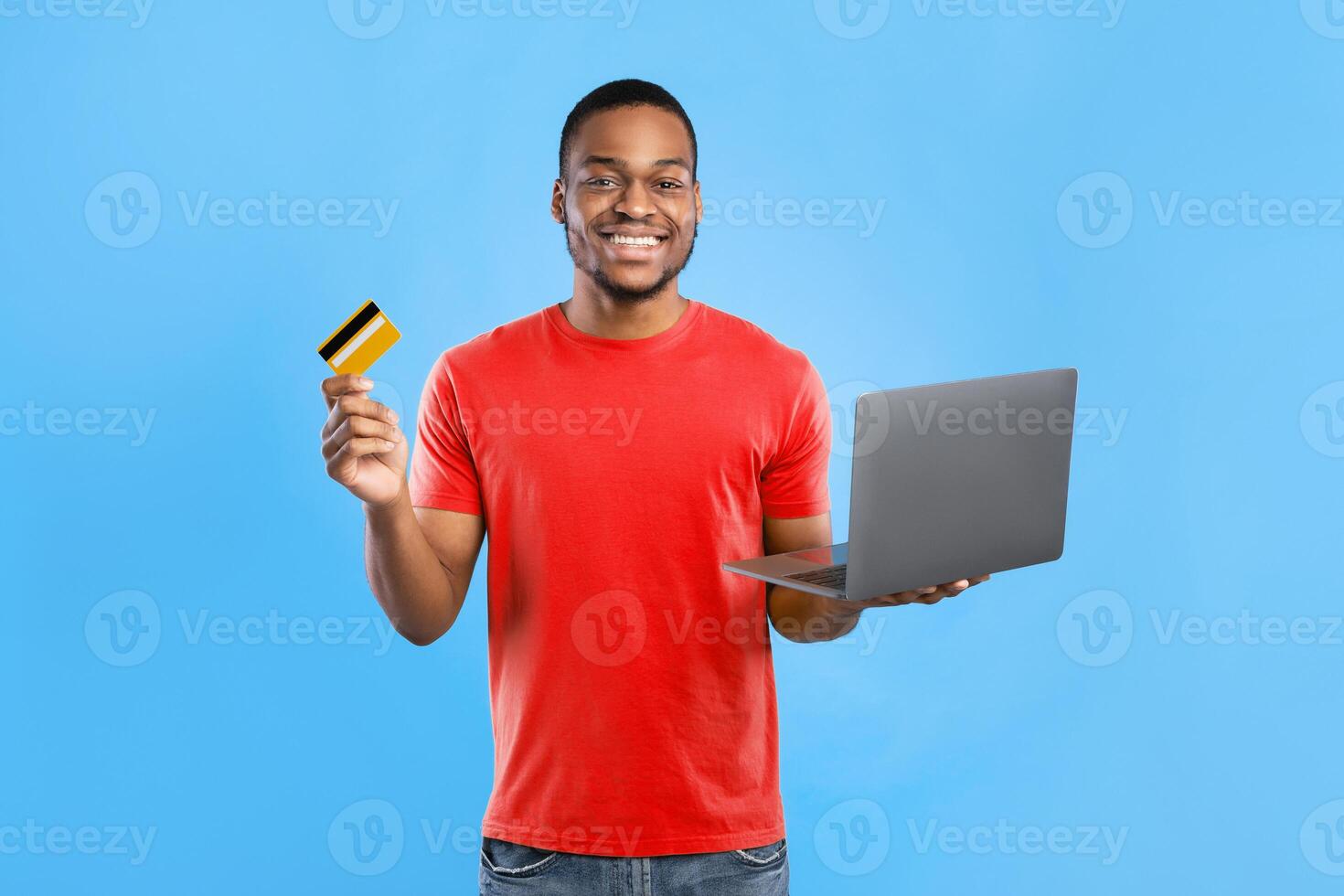 Online Shopping Concept. Black Man Holding Laptop And Credit Card Smling To Camera Standing Over Blue Background In Studio. Customer Buying Something In Internet Store photo