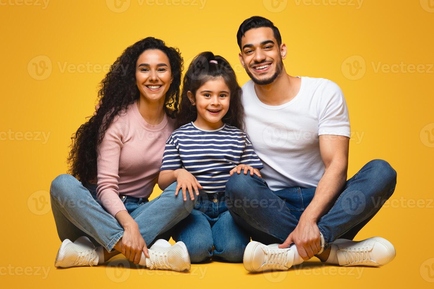 Portrait Of Happy Young Middle Eastern Family Of Three With Little Daughter. Cheerful Arab Parents And Their Cute Female Kid Sitting On Floor Over Yellow Studio Background And Smiling At Camera photo