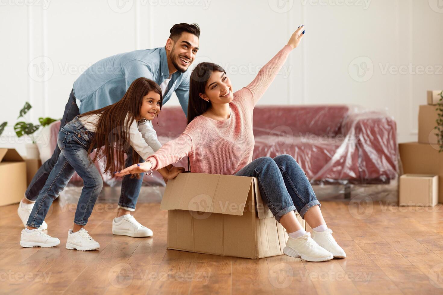 Happy family playing in new own home, buyers have fun, laughing on moving day, excited guy riding lady sitting in cardboard box, husband and daughter pushing carton package. Relocation, rent and break photo