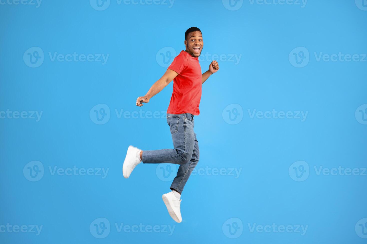 Carefree african american young man jumping smiling to camera in studio on blue background. Full length shot of happy guy posing in mid-air. Positive emotions and happiness photo