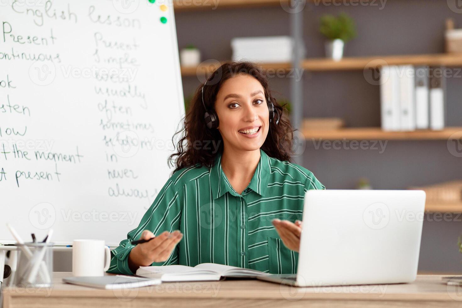 Distant Learning Concept. Portrait of smiling teacher talking and gesturing to camera wearing wireless headphones sitting at desk with pc laptop near whiteboard. Lady explaining educational material photo