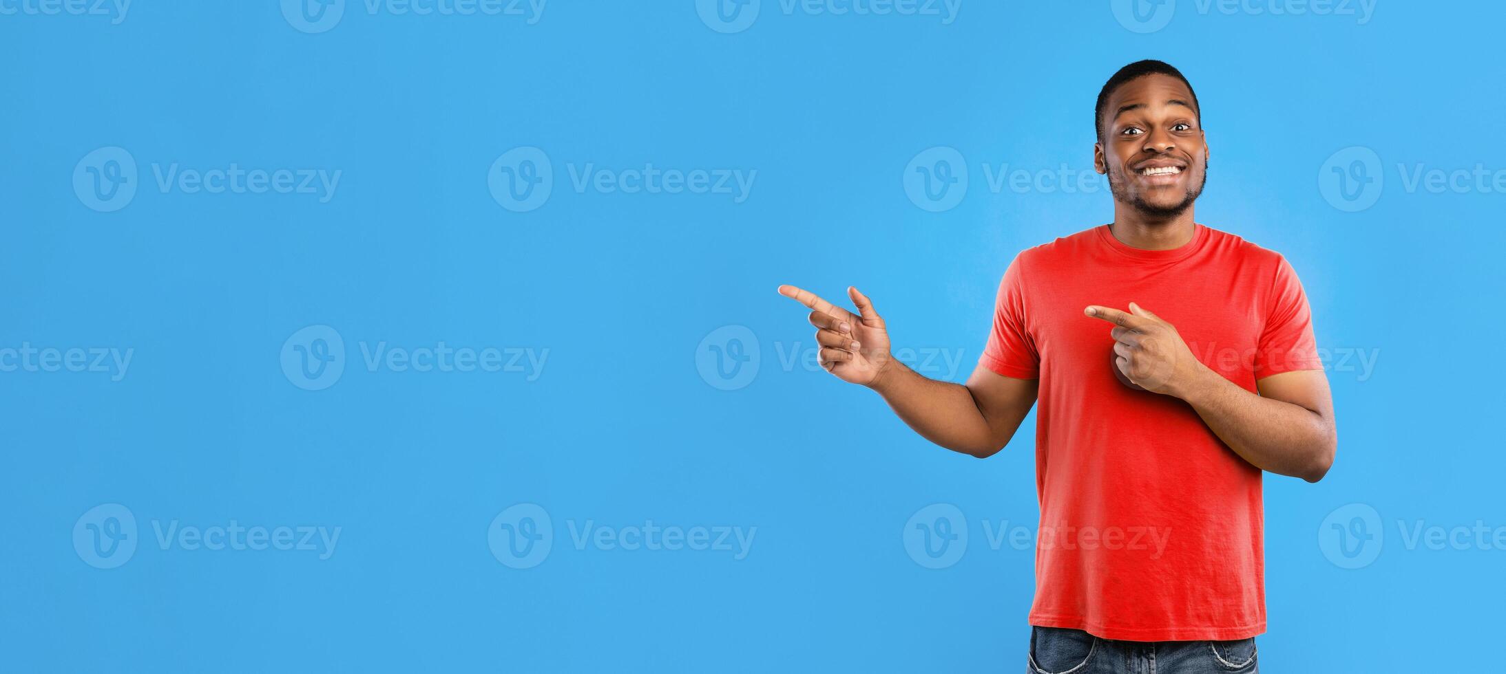 Look Aside. Black Male Pointing Fingers Showing Copy Space For Advertisement Text Standing Wearing Yellow T-Shirt On Blue Studio Background, Smiling To Camera. Panorama photo