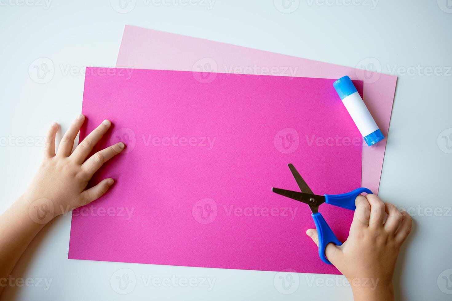 Childs hands crafting with pink paper, blue scissors, and glue on light background photo