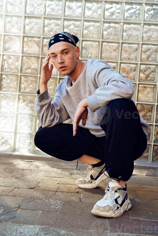 A young man wearing a bandana sits with his legs crossed in front of a glass block wall photo