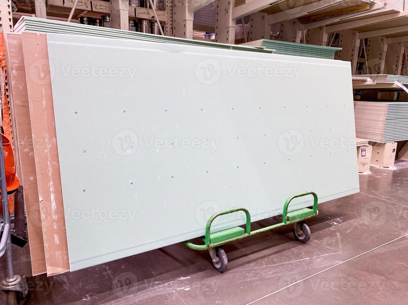 A green cart is currently transporting a sizable piece of drywall that is necessary for construction projects and renovations in various settings, such as buildings and offices photo