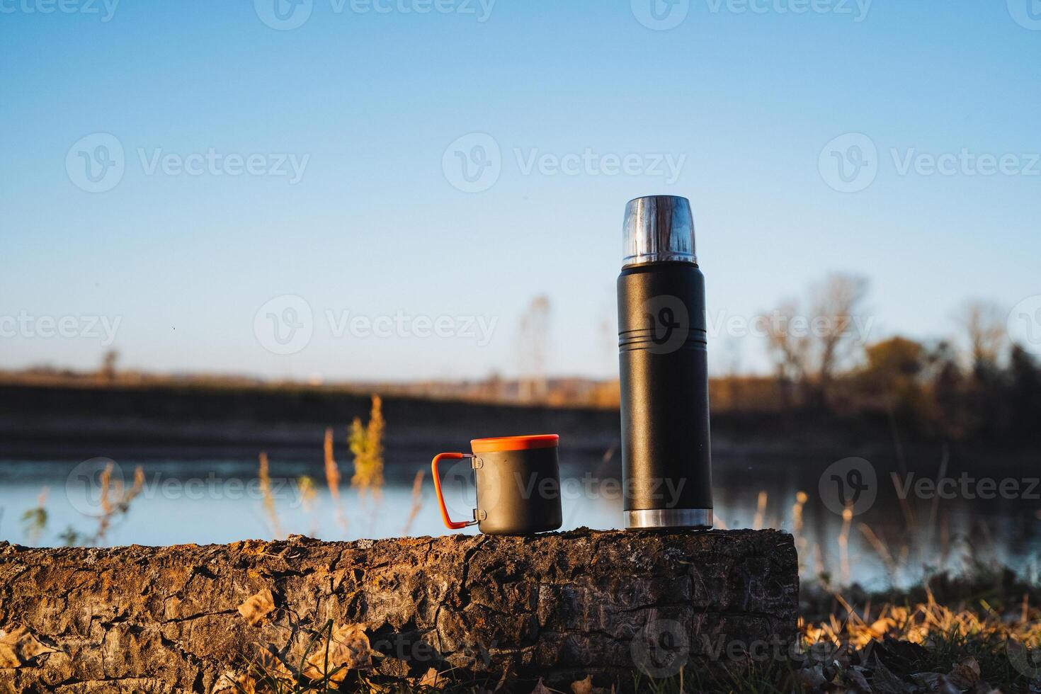 A thermos and a cup are sitting on a rock near a lake photo