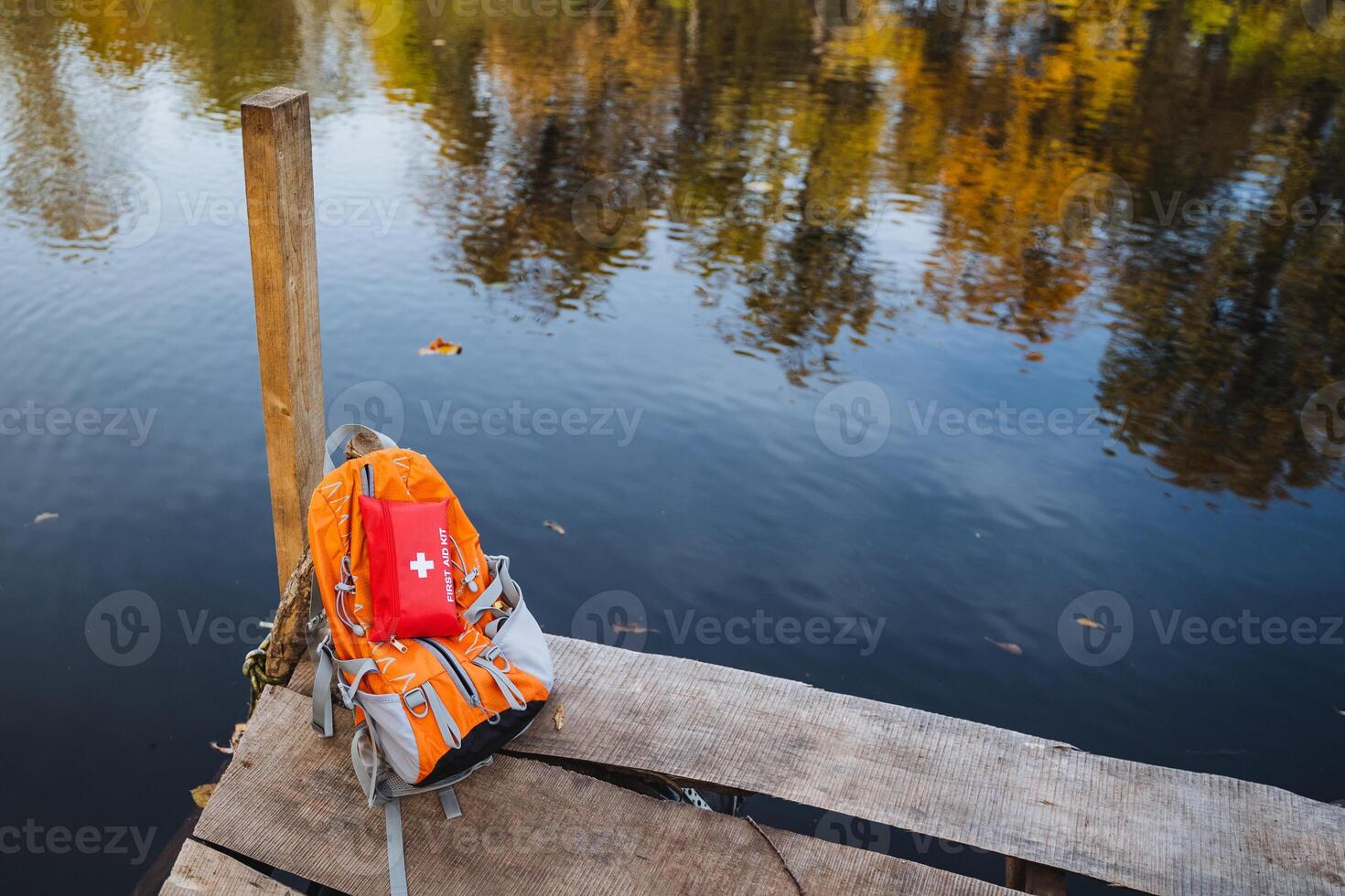 A serene autumn scene features a vibrant backpack resting by calm, reflective waters, creating a perfect setting for exploration and relaxation photo