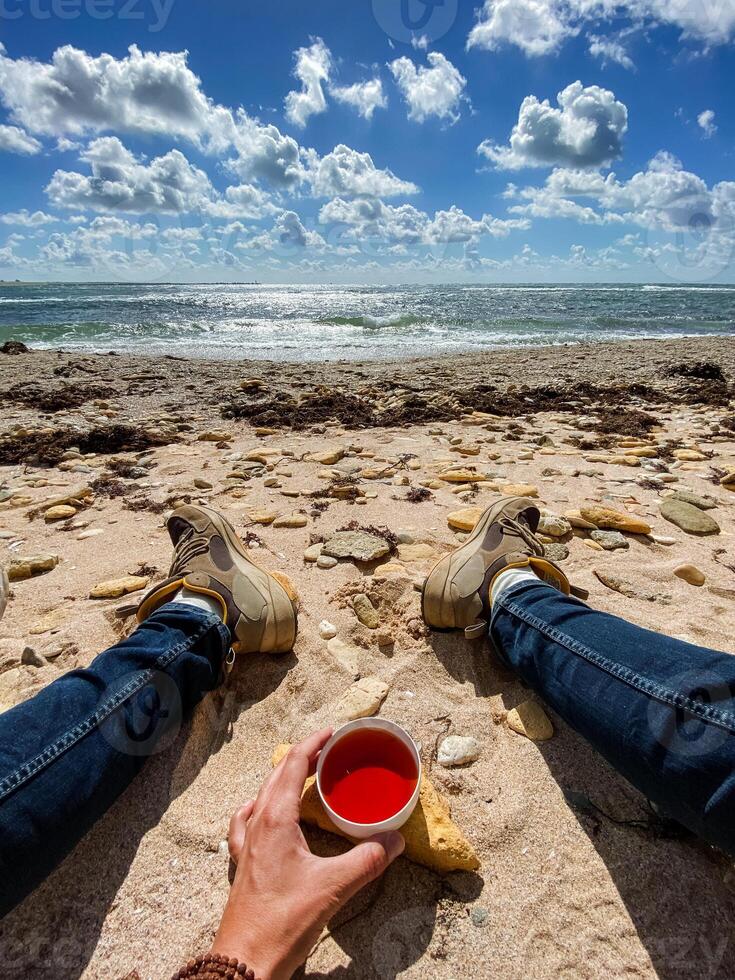 A person is leisurely sitting on the beach while holding a cup filled with a refreshing red liquid, enjoying the wonderful ambiance of summer by the seaside, with the sun shining brightly photo