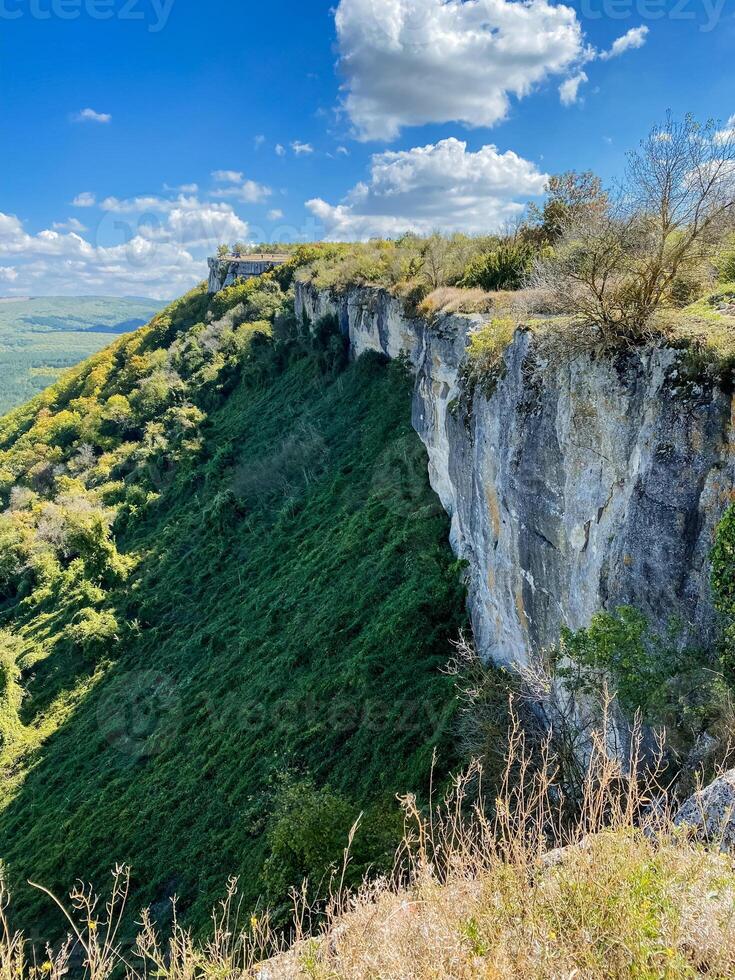 There is a towering cliff adorned with lush green trees, standing majestically against a backdrop of a clear blue sky that extends infinitely in all directions, inviting serenity and beauty photo