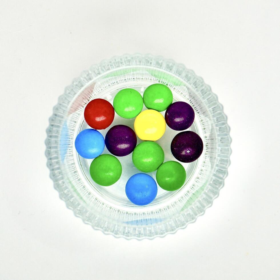 Group of colorful small round rainbow chocolate candy in round glass bowl isolated on square white background from top angle view. photo