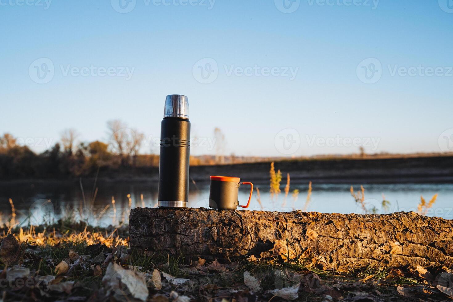 A classic thermos and a ceramic cup sit gracefully together on a smooth rock near a tranquil lake, surrounded by breathtaking scenery, beneath a clear blue sky and gentle, lapping waves photo