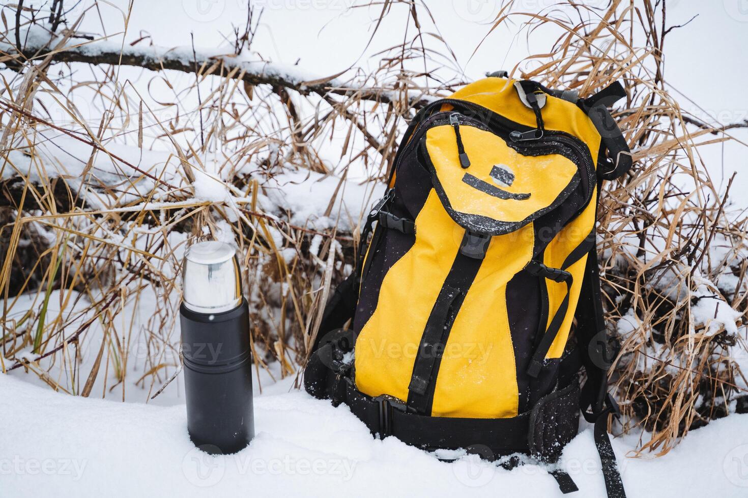 In the serene winter landscape, a vibrant yellow backpack rests in the soft white snow, accompanied by a sturdy black thermos, creating a striking and beautiful contrast photo