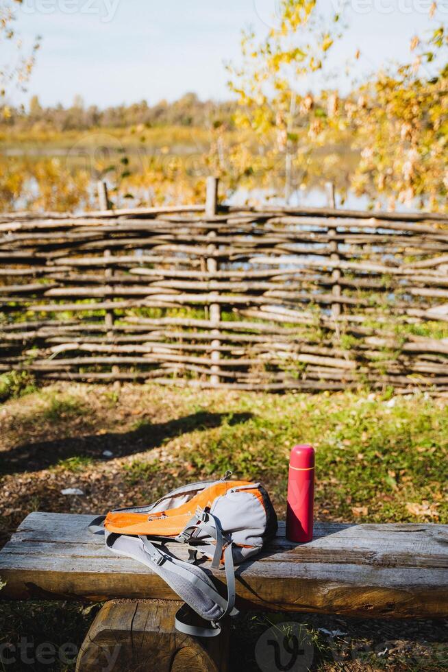A backpack along with a thermos is resting on a rustic wooden bench, surrounded by the beauty of nature and rural landscapes, highlighting the charm of the outdoor environment photo