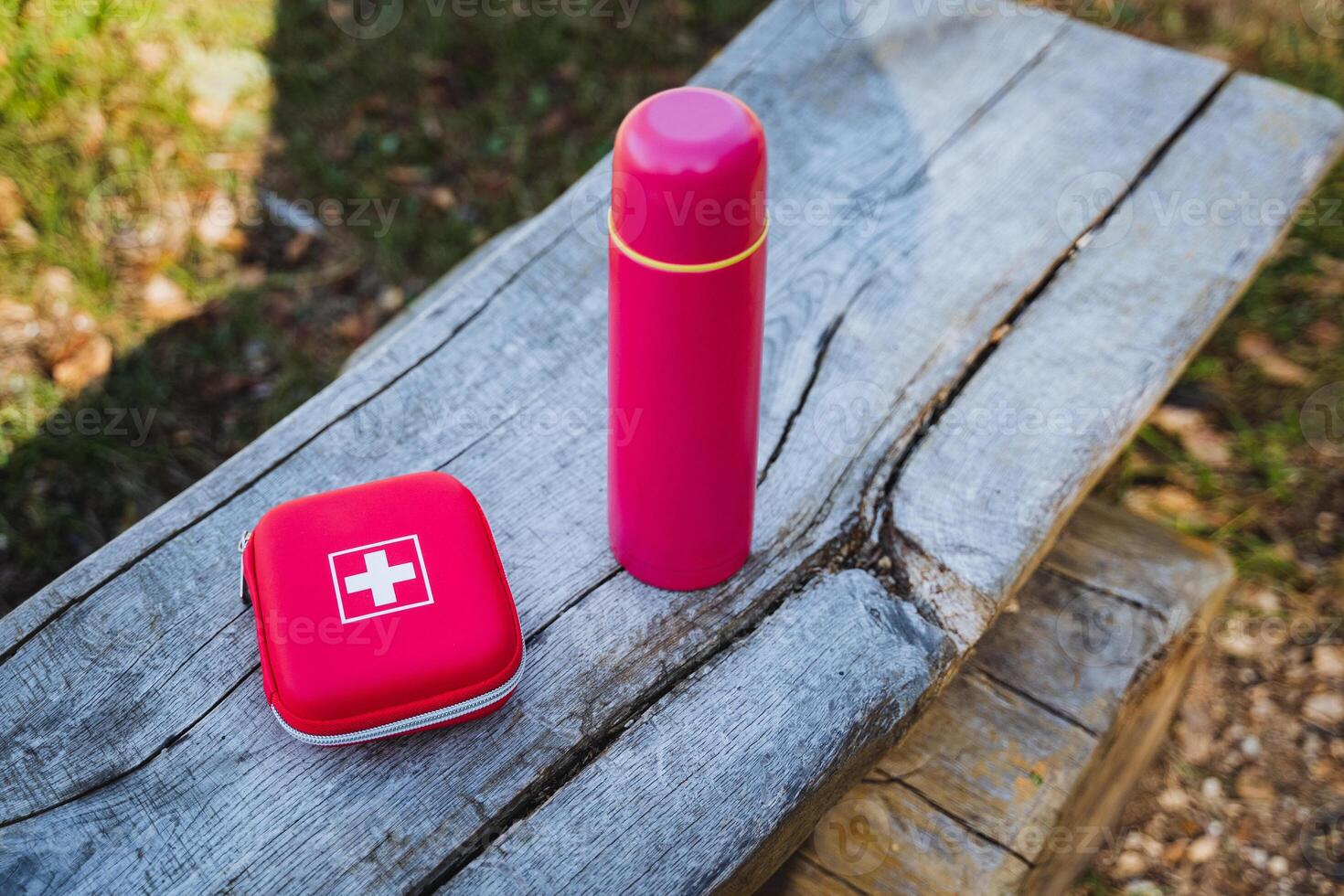 A charming pink thermos, accompanied by a bright red first aid kit, can be discovered resting on a rustic wooden bench that is elegantly situated in a tranquil and peaceful environment photo