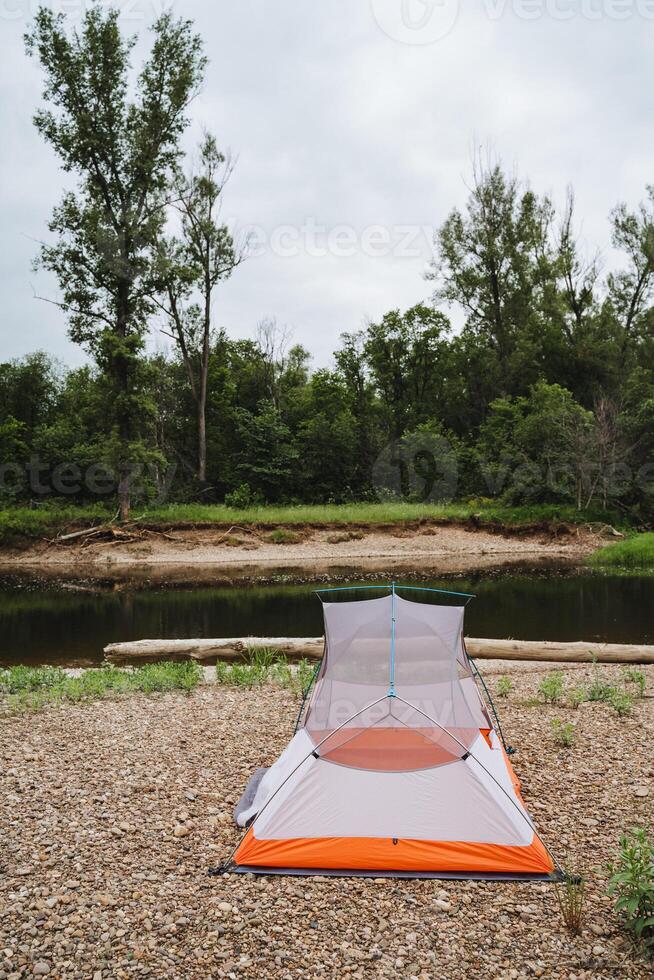 Imagine the tranquility of camping next to a calm riverside in nature, surrounded by greenery with a tent on a pebble shoreline, offering relaxation and a scenic view photo