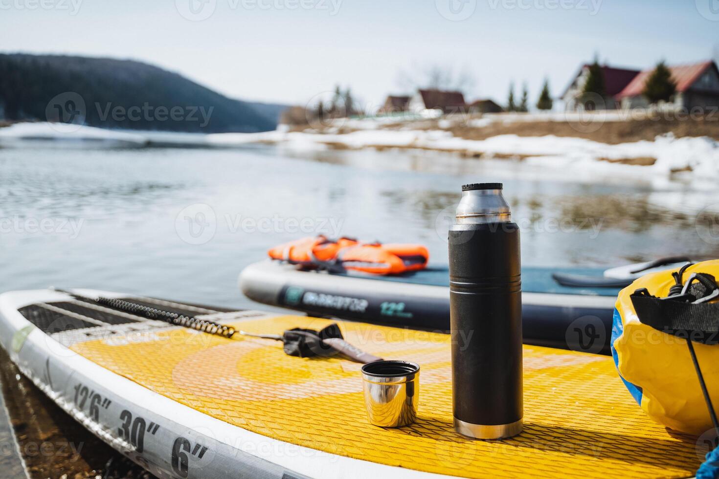 A thermos with a mug stands on a SUP against the background of a water landscape, a hot drink in cold weather, a tourist rafting on a SUP board. photo