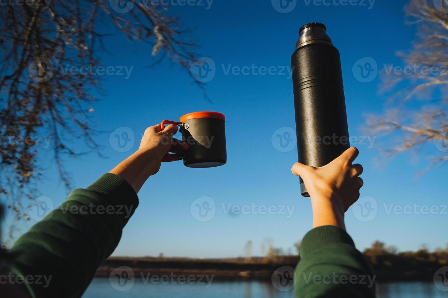 A person is seen holding a cup filled with a warm beverage in one hand, while clutching a thermos of a hot drink in the other, savoring a beautiful day outdoors, immersed in nature photo