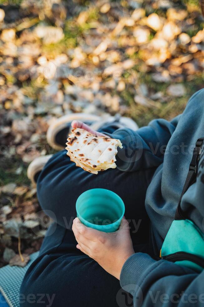 A person is comfortably sitting on the ground, enjoying a delicious sandwich while simultaneously sipping on a warm cup of coffee, taking in the surrounding scenery of nature photo