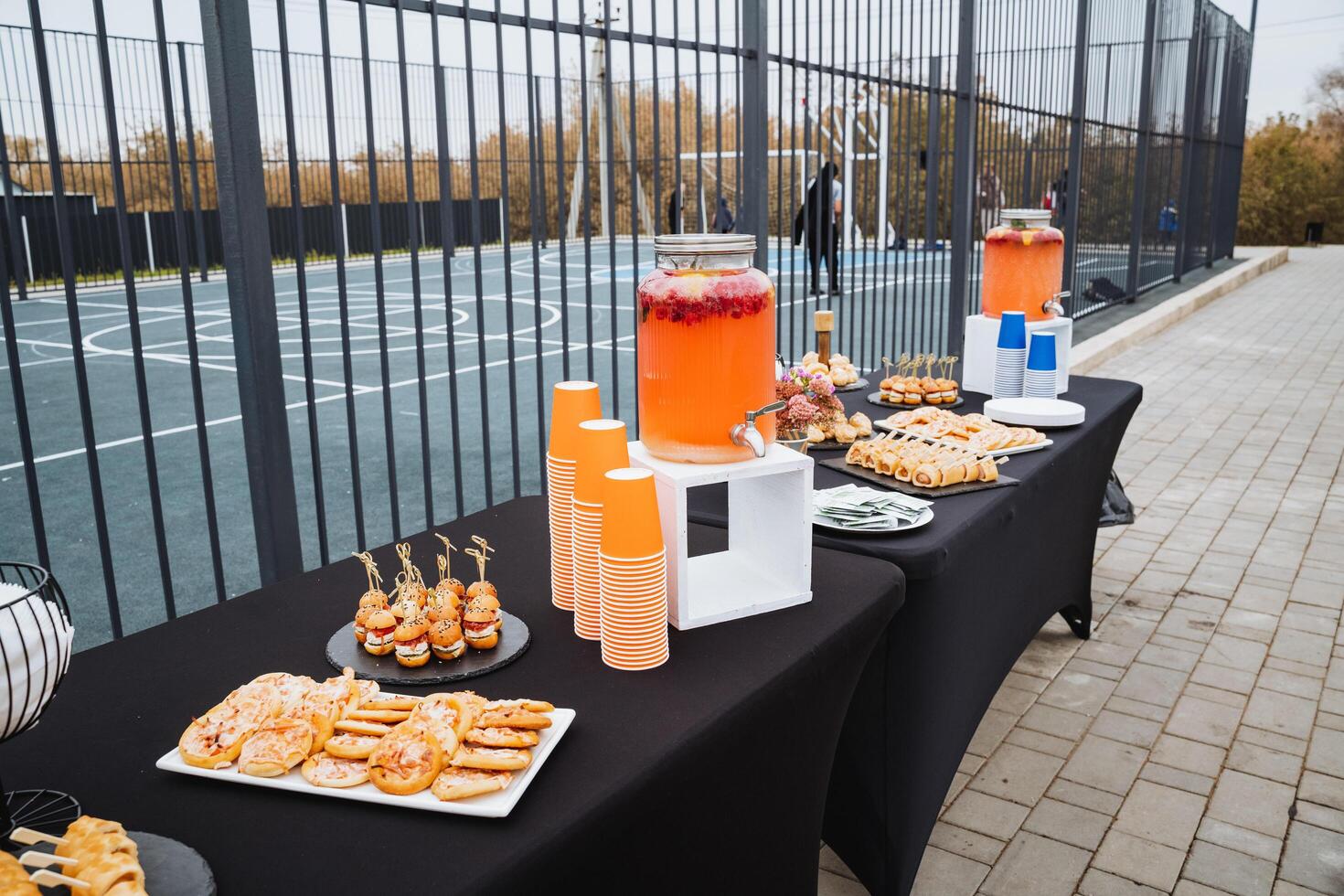 There is a nicely arranged table adorned with various delicious food items and refreshing drinks situated right in front of a wellmaintained tennis court, creating a perfect setting photo