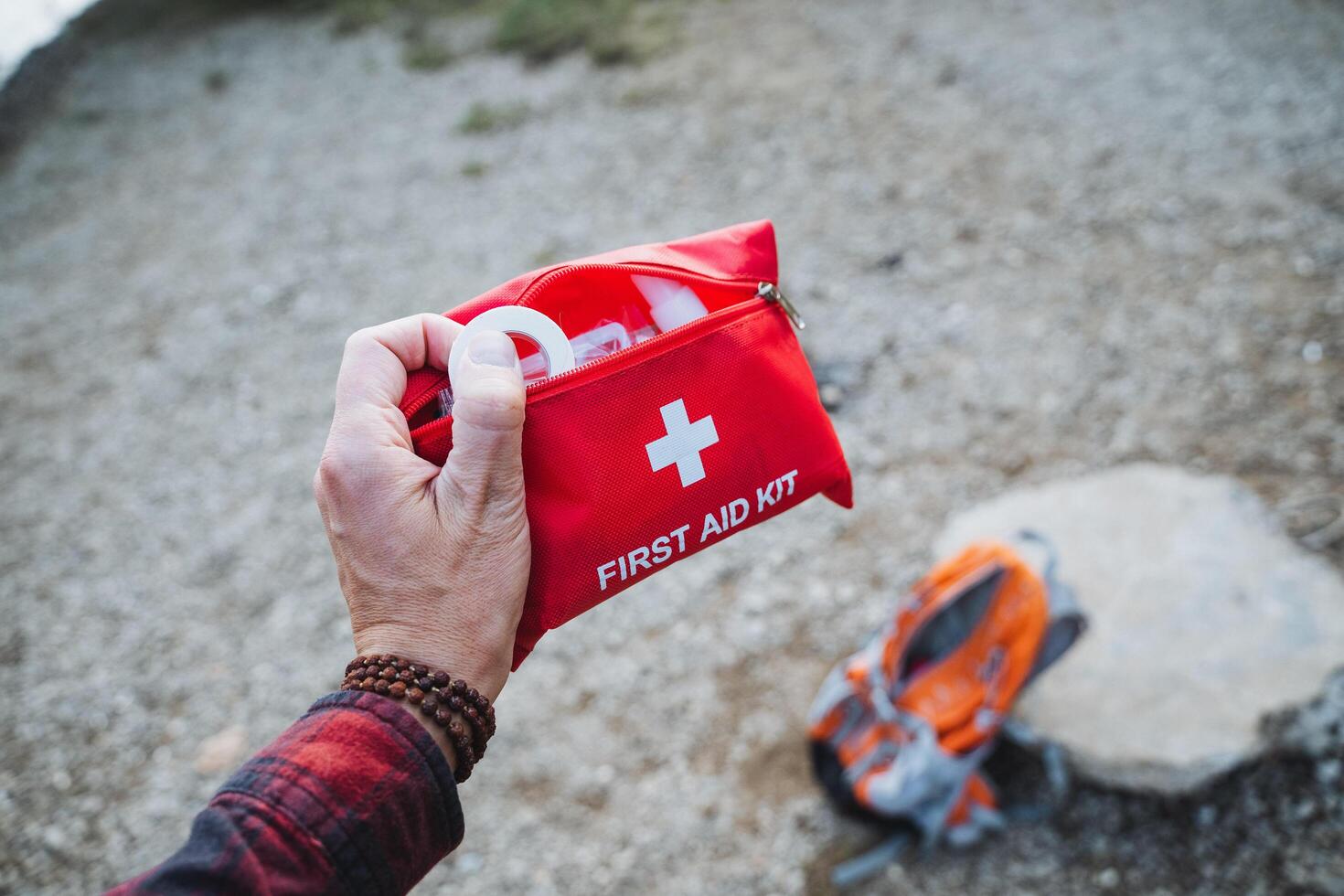 A bright red First Aid Kit is held firmly in hand while a sturdy backpack rests securely on the ground, ensuring outdoor safety and preparedness photo