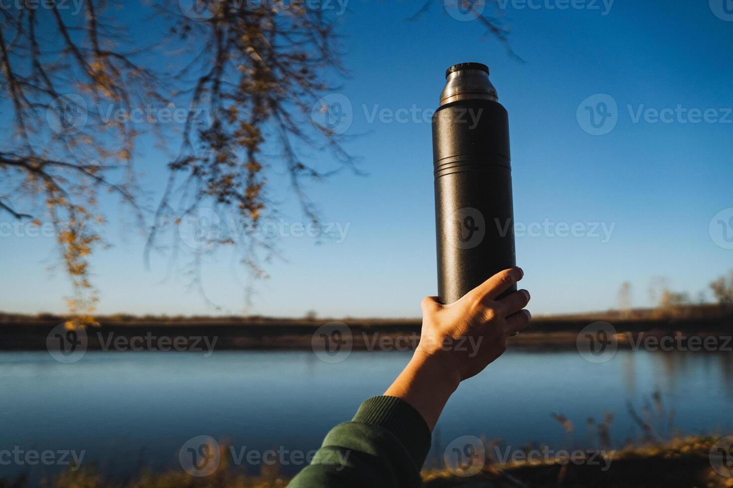 A person stands by a serene lake, holding a thermos in their hand, gazing over the calm water, surrounded by lush plants and trees under a beautiful sky filled with clouds photo