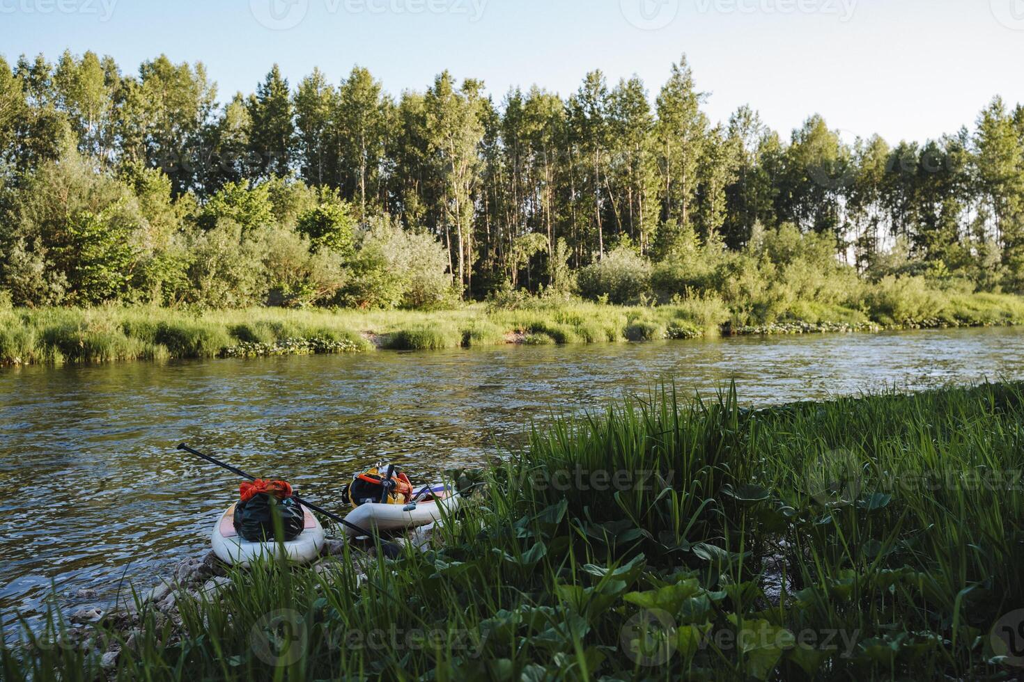 The serene riverbank features lush greenery, tall trees, and an inflatable boat, perfect for outdoor adventures and relaxation in natures beauty and tranquility photo