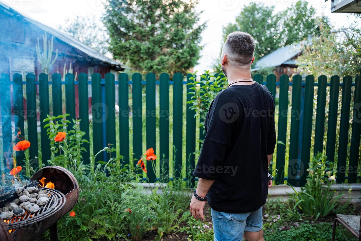 A man is grilling at a backyard BBQ surrounded by blooming flowers and a fence, enjoying the summer outdoors with friends and family in a relaxed and casual setting photo