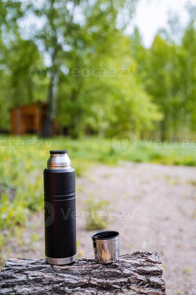 A glass bottle and a cup of drink are resting on a tree stump in the woods. Surrounded by grass and plants, they contrast with the natural wood and asphalt road surface nearby photo