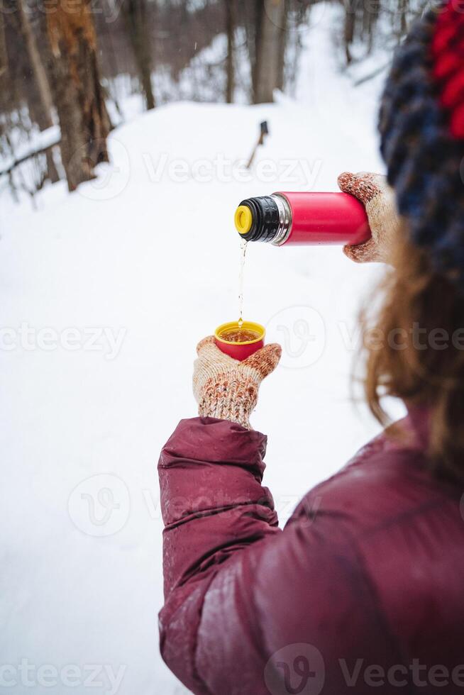 A woman is carefully pouring steaming coffee from a thermos into a cup while standing in the snow, surrounded by a winter wonderland of white, chilly flakes falling gently around her photo
