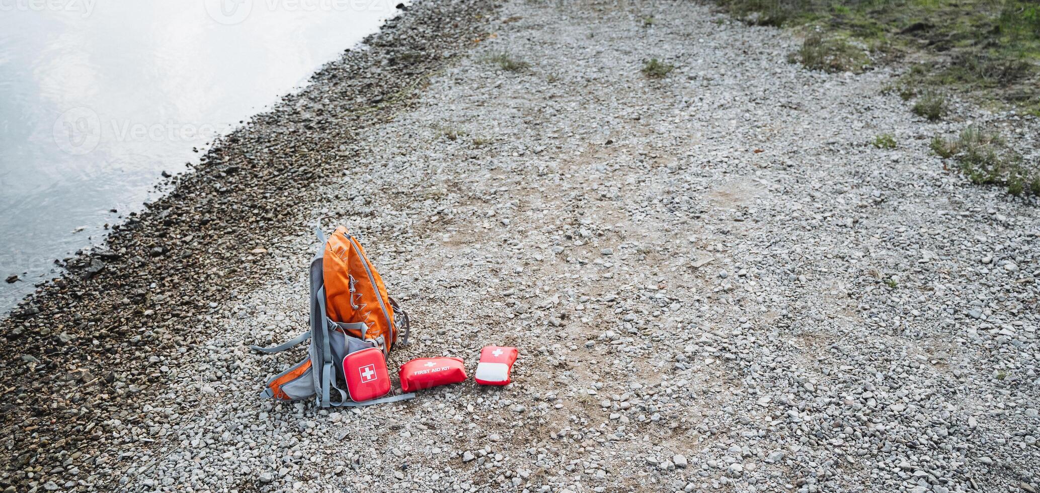 A backpack and a first aid kit can be seen laying on the ground next to the edge of a body of water, possibly indicating someone was preparing for an outdoor adventure nearby photo