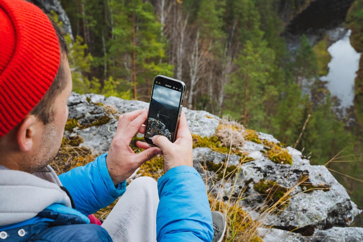A man, comfortably relaxed on a large rock amidst the wilderness, captures and preserves the stunning beauty of nature surrounding him using his cell phone to remember this moment forever photo