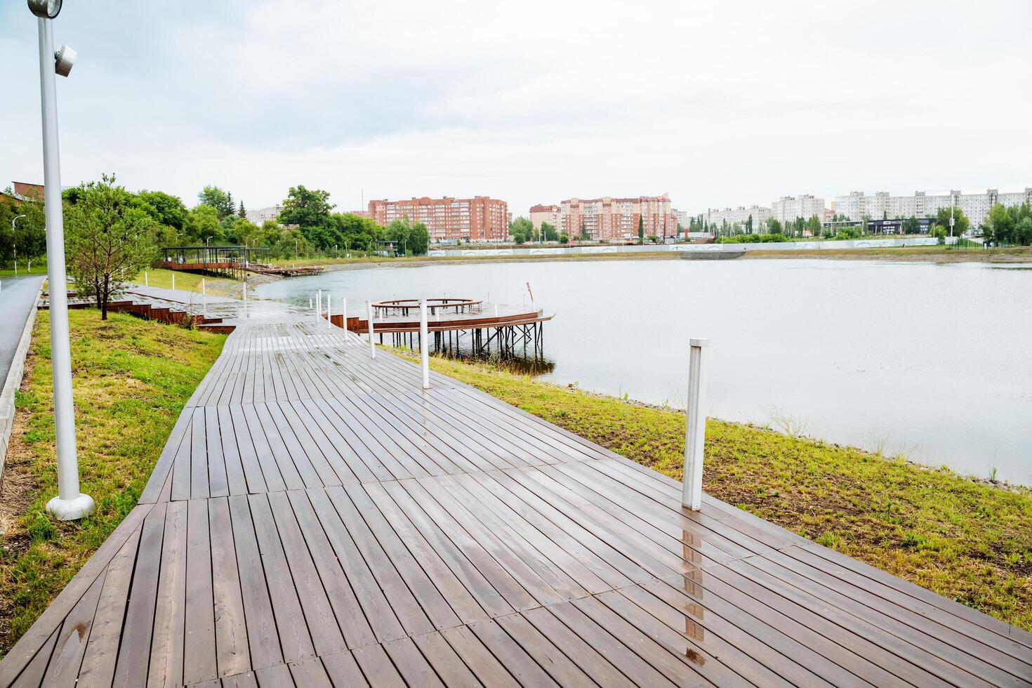 A serene park features a wooden pathway leading to a calm lake, integrating natural beauty with manmade elements. The scene highlights the harmony between nature and human design photo