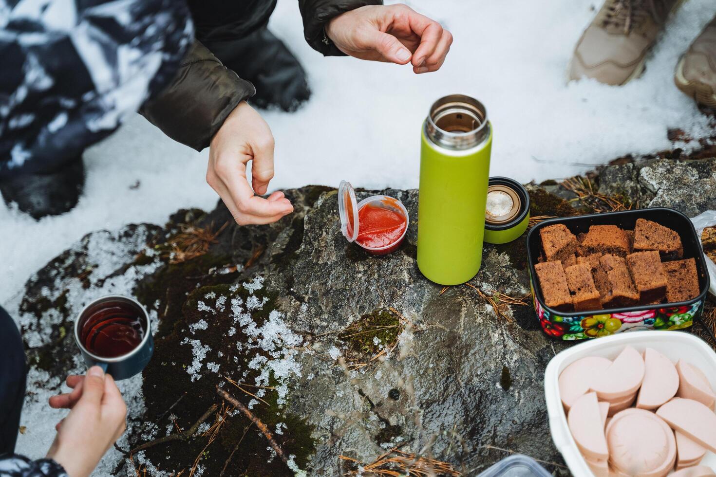 Food on a hike in winter, a thermos with tea stands on a stone, sausage lies in a container, a mug of tea, top view of a picnic in nature. photo